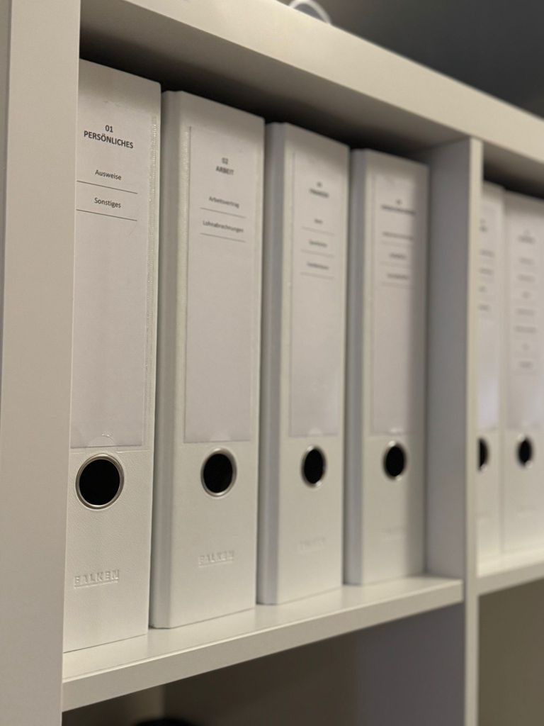 Weiße Aktenordner mit einheitlichen Rücken-Beschriftungen wie 01 Persönliches und 02 Arbeit in einem Regal